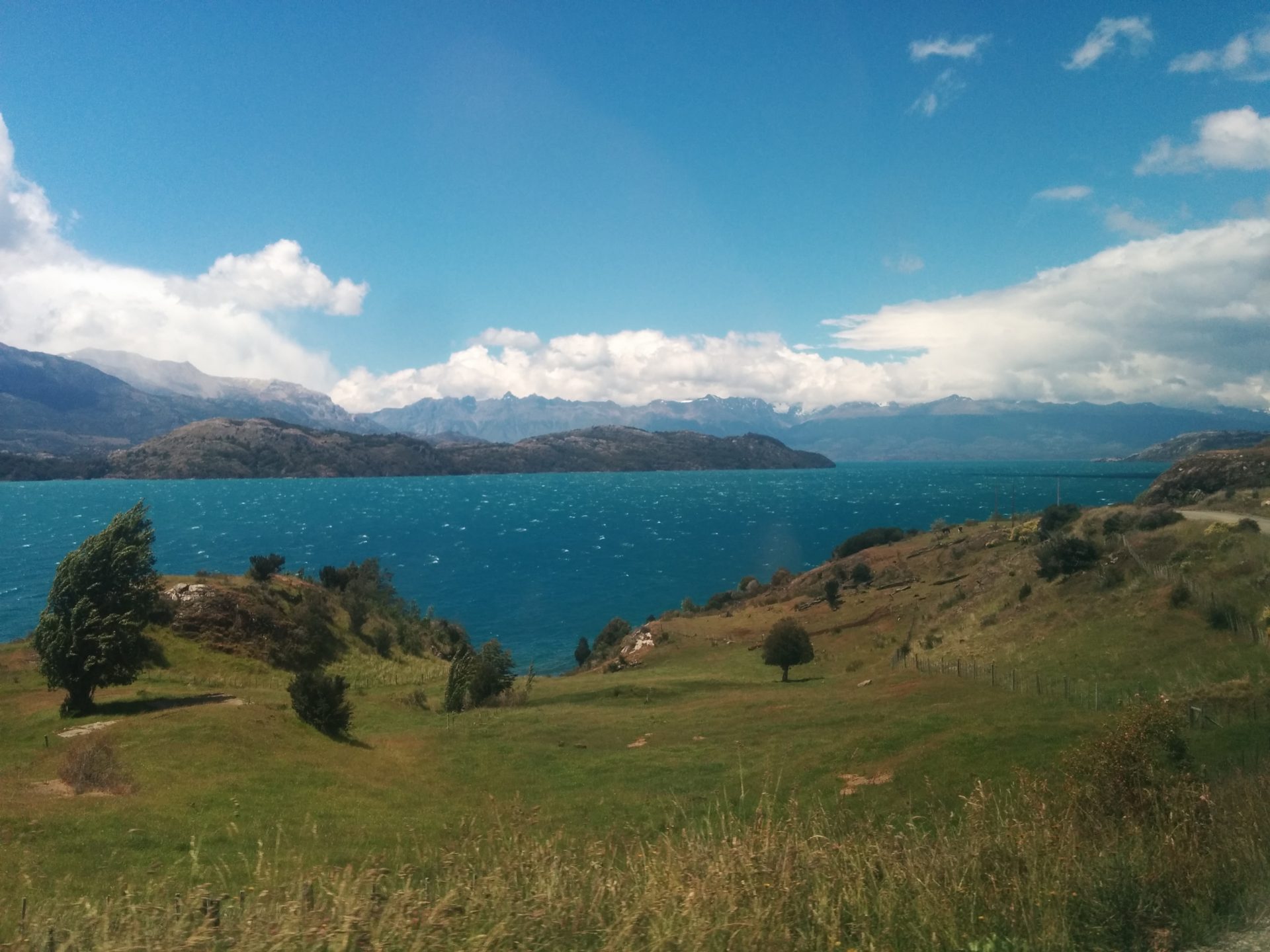 A turquoise lake behind a green field and in front of mountains.