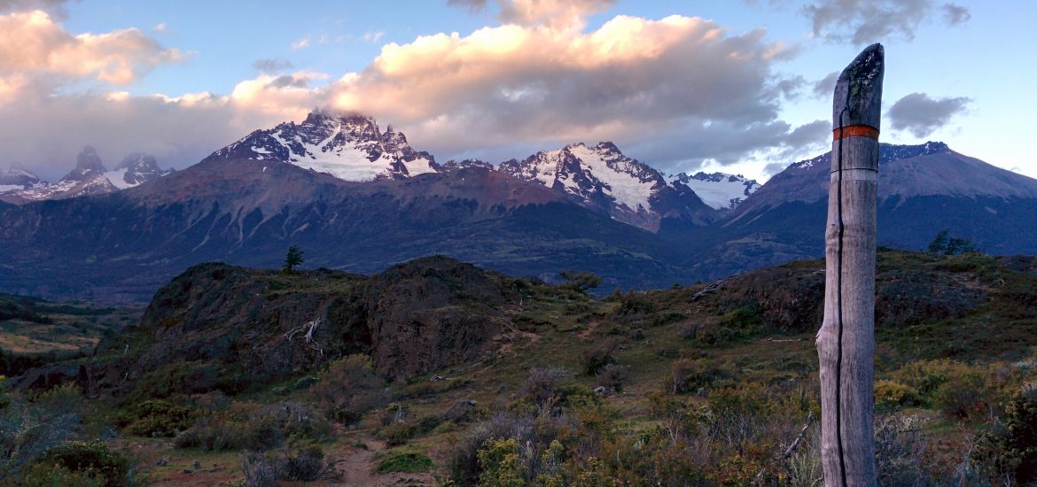 In the nature during a sunset, the Cerro Castillo in the background. There is a signpost that shows the way.