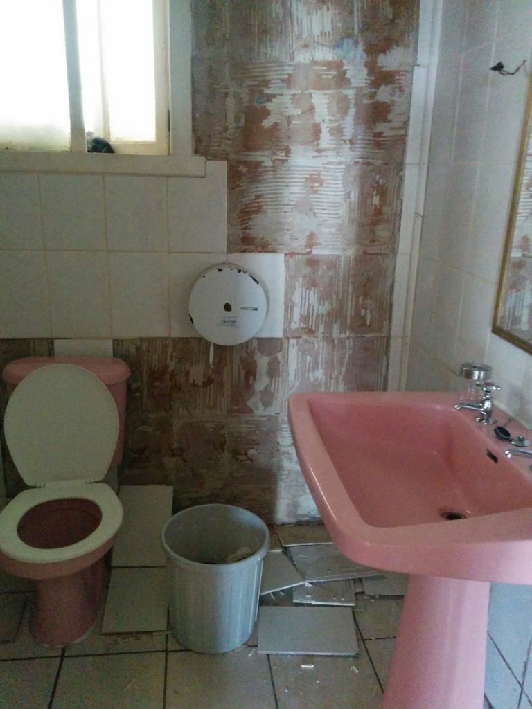 A bathroom where the tiling is off the wall and lying broken on the ground.